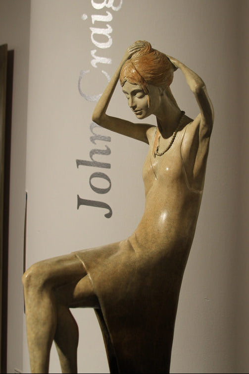 A sculptural nude female figure in warm tan and brown tones, posed with arms raised, displayed against a gallery wall with vertical text reading 'John Craig Brown'.